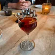 A red-brown cocktail with a skewered cherry sits in a stemmed glass on a marble table.