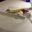 A slice of cheesecake on a rectangular plate with whipped cream, a strawberry, sauce, and a dusting of brown powder.