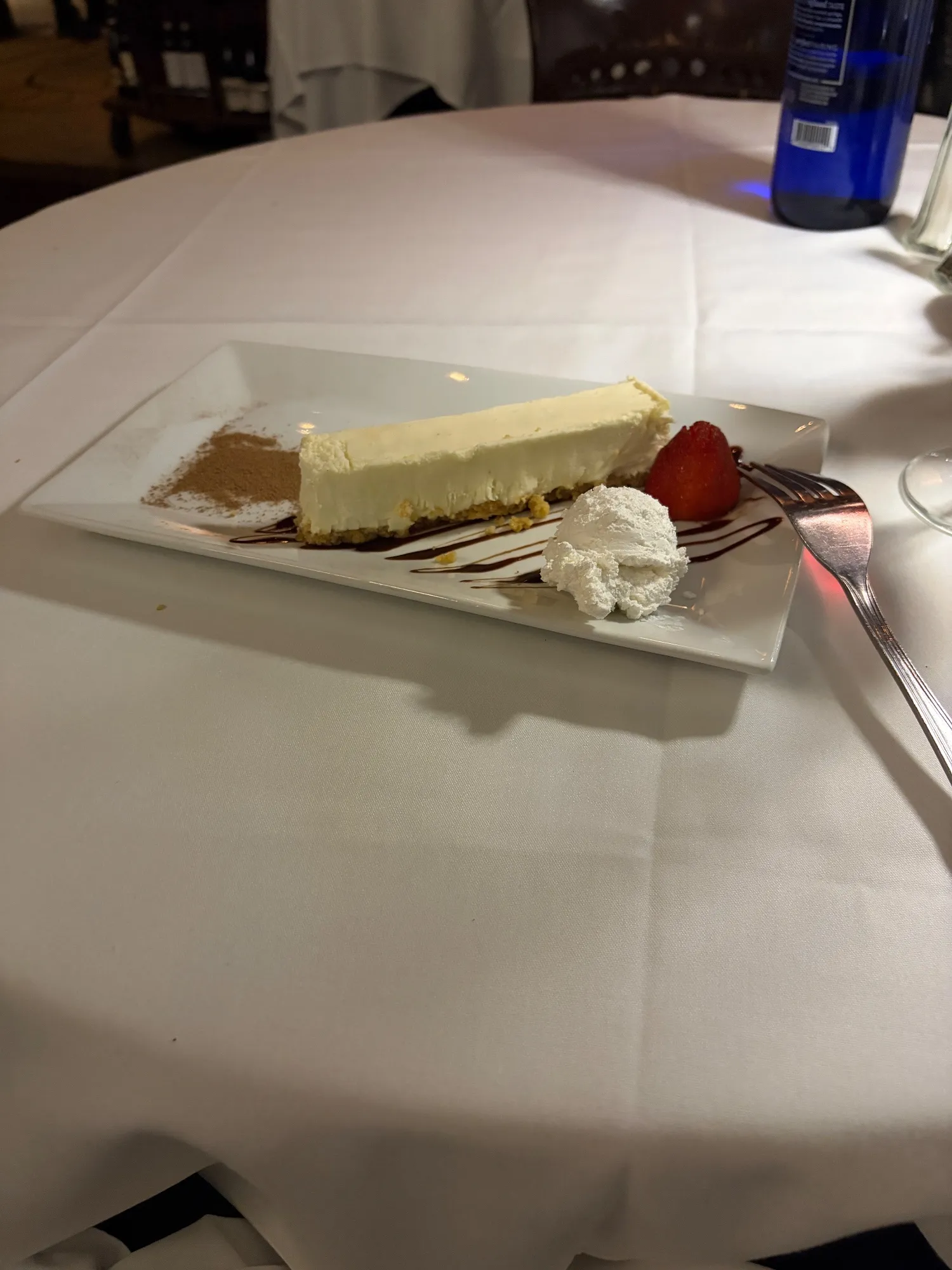 A slice of cheesecake on a rectangular plate with whipped cream, a strawberry, sauce, and a dusting of brown powder.