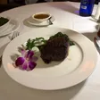 A filet mignon on a white plate with greens, parsley, and a purple flower, with a sauce boat behind it.