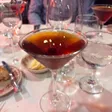 A dark cocktail in a martini glass with a cherry, on a white tablecloth among wine glasses.