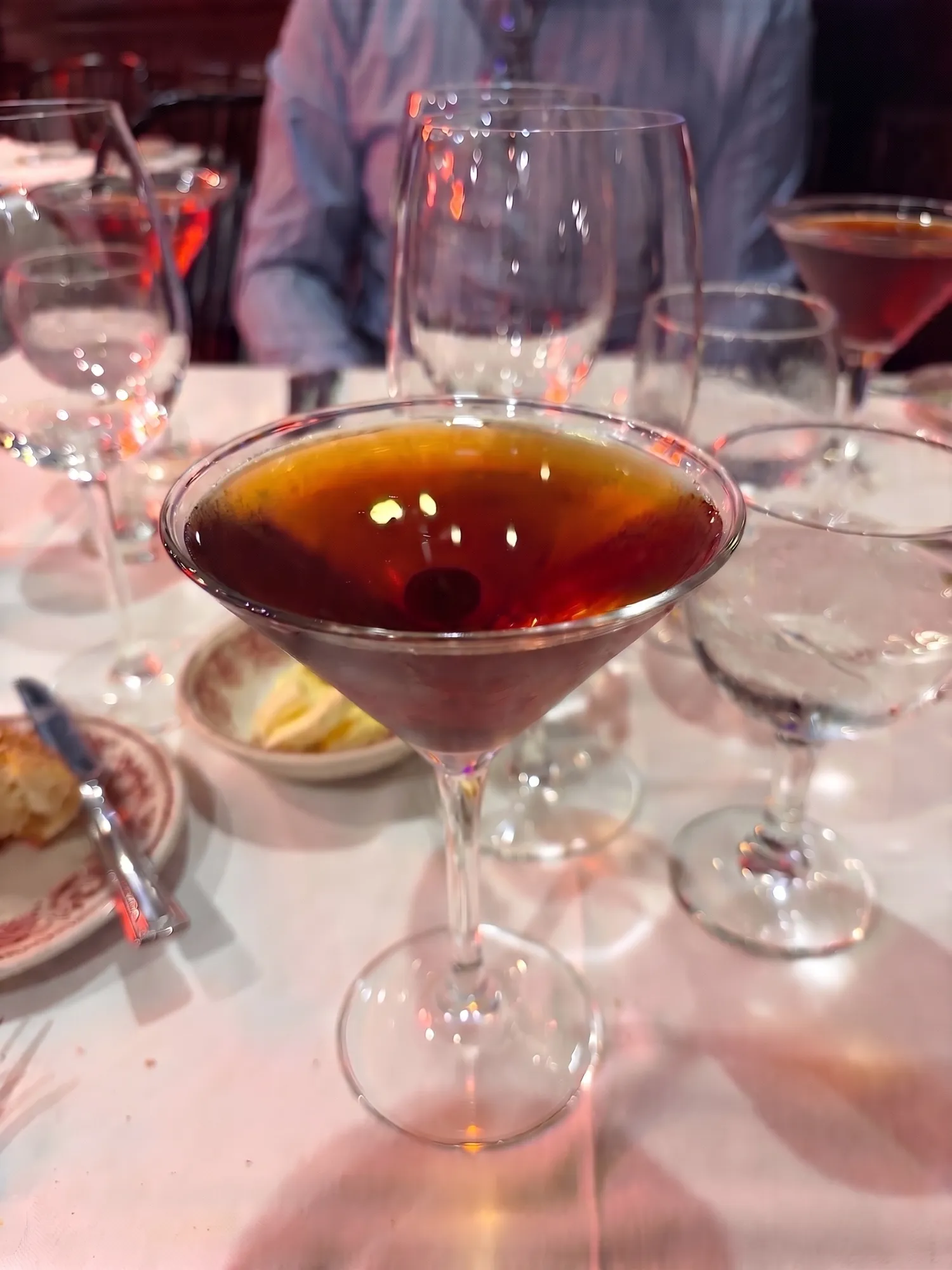 A dark cocktail in a martini glass with a cherry, on a white tablecloth among wine glasses.