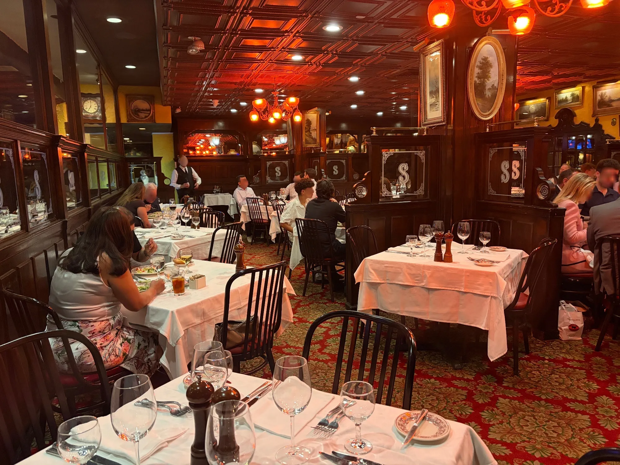 Dining room with wood paneling, framed paintings, patterned red carpet and warm overhead lighting, tables set with white cloths.