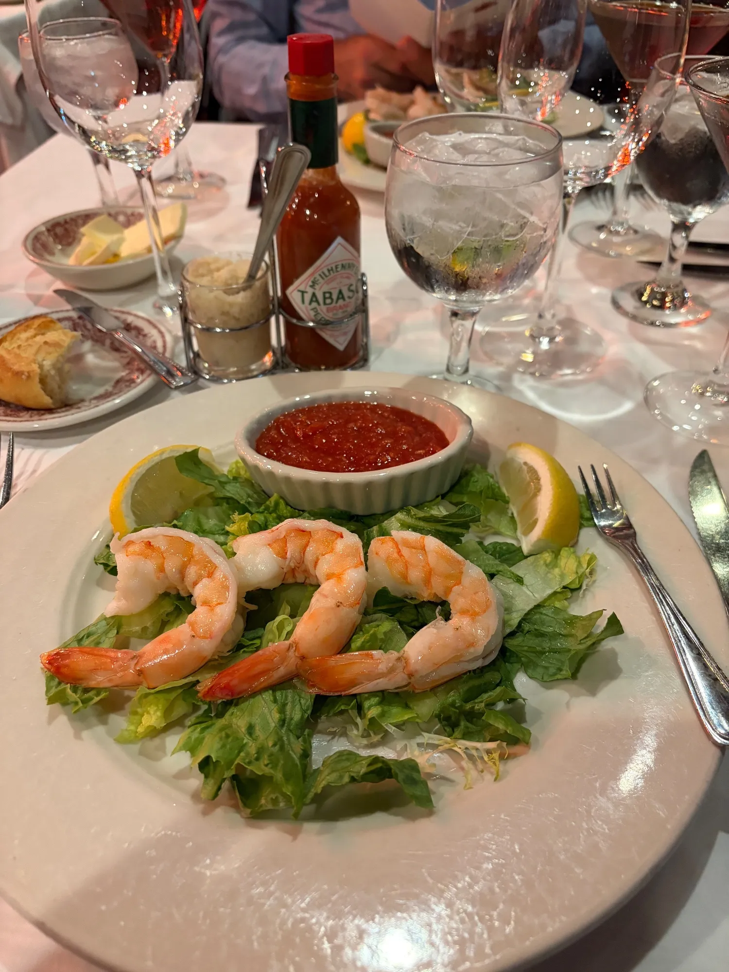 Five shrimp arranged on lettuce around a ramekin of cocktail sauce, with lemon wedges, a Tabasco bottle and horseradish nearby.