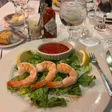 Five shrimp arranged on lettuce around a ramekin of cocktail sauce, with lemon wedges, a Tabasco bottle and horseradish nearby.