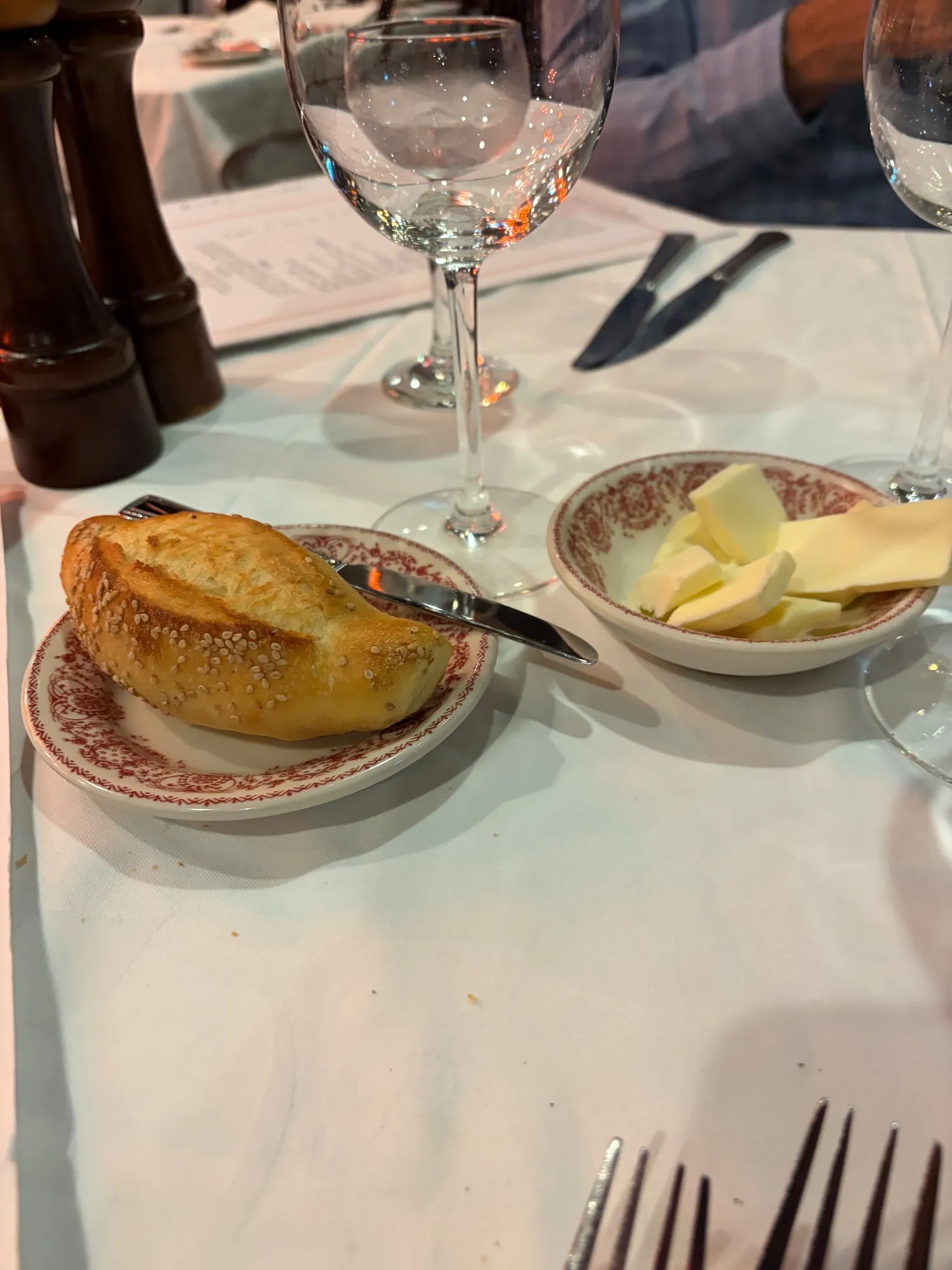 A roll of bread with sesame on a plate with a knife, beside a dish of butter and wine glasses.
