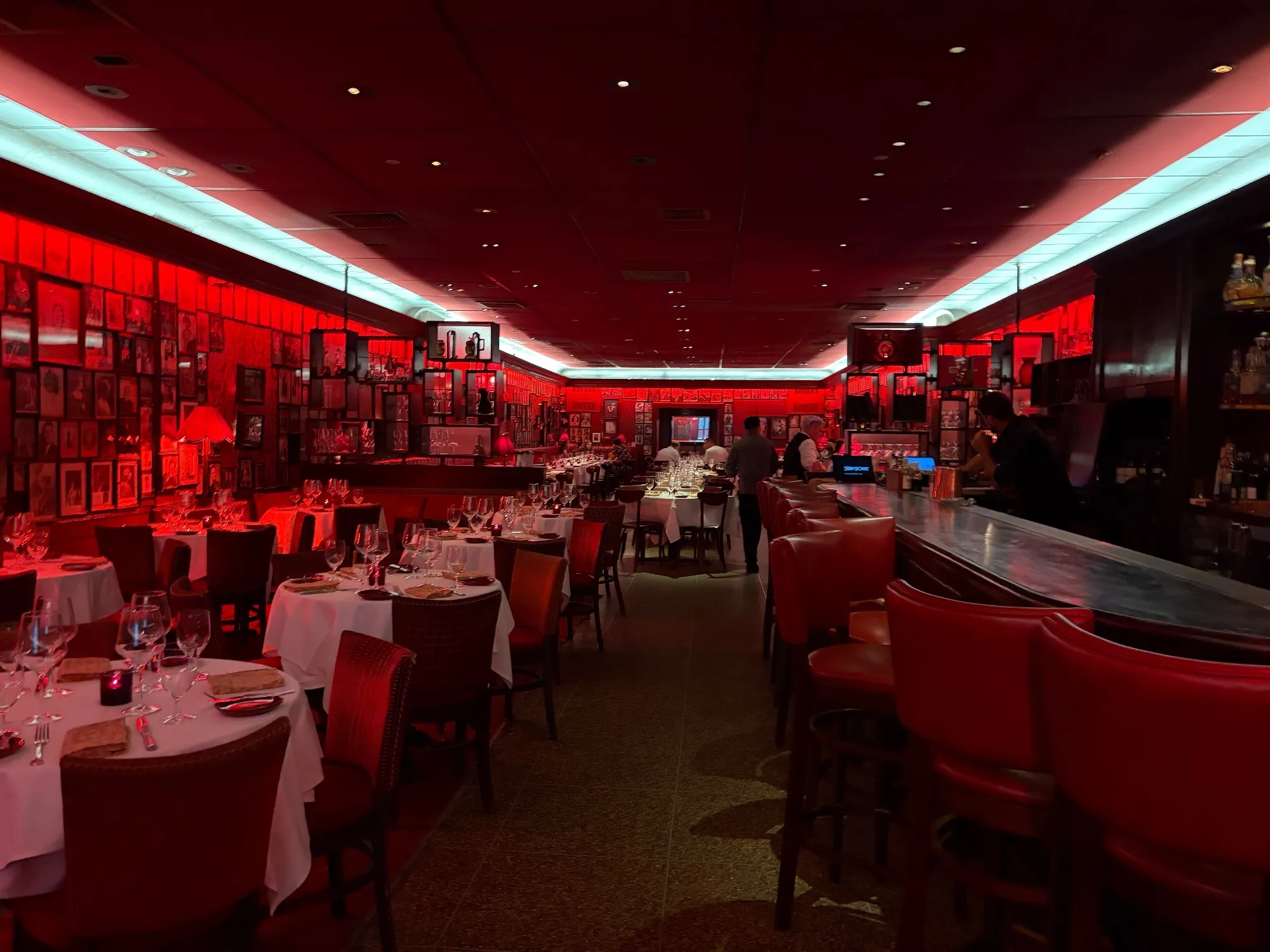 A red-lit dining room with white-tablecloth tables, framed pictures covering the walls, and a bar along one side.