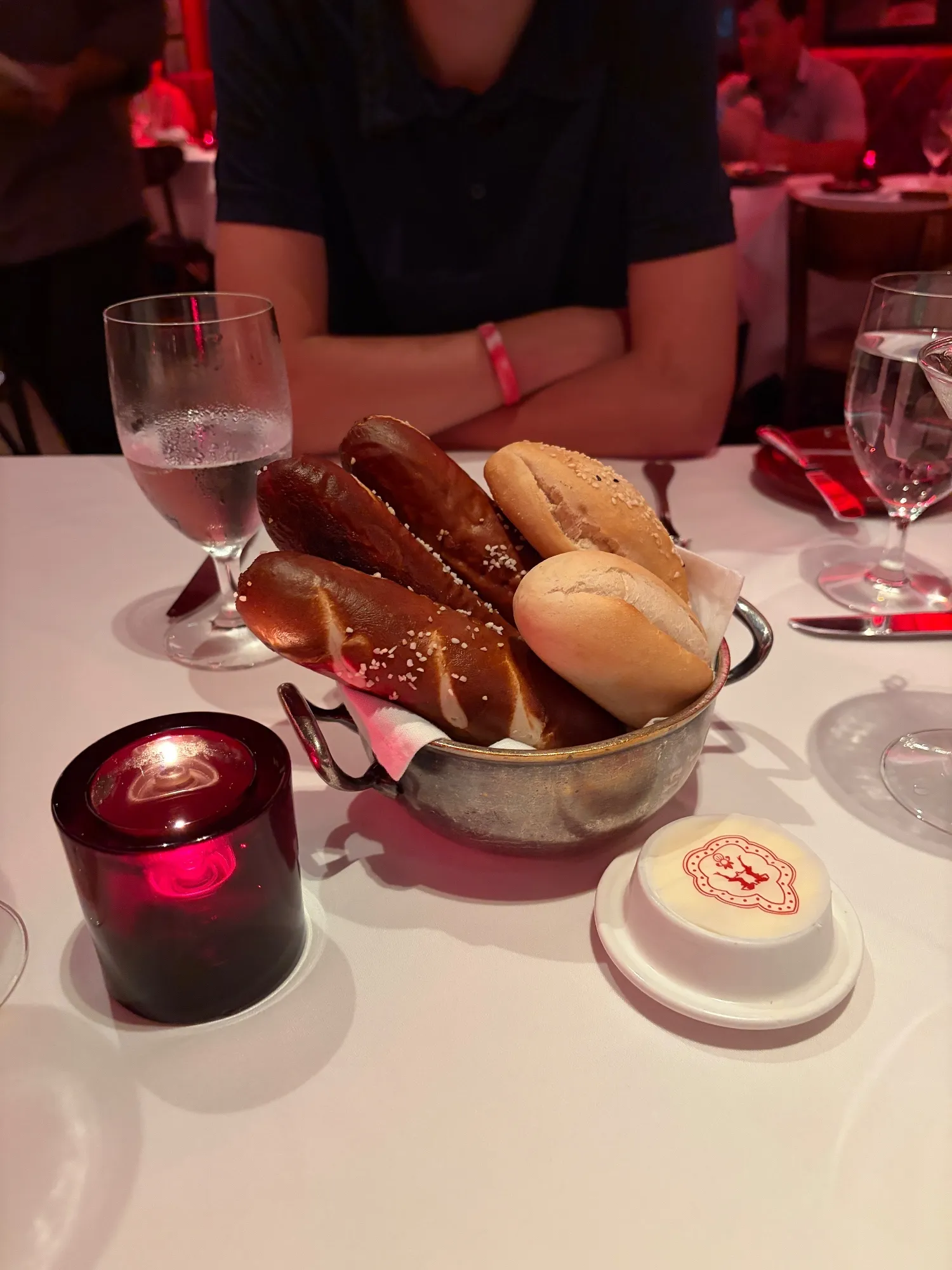 A metal basket containing pretzel bread and rolls beside a covered butter dish.