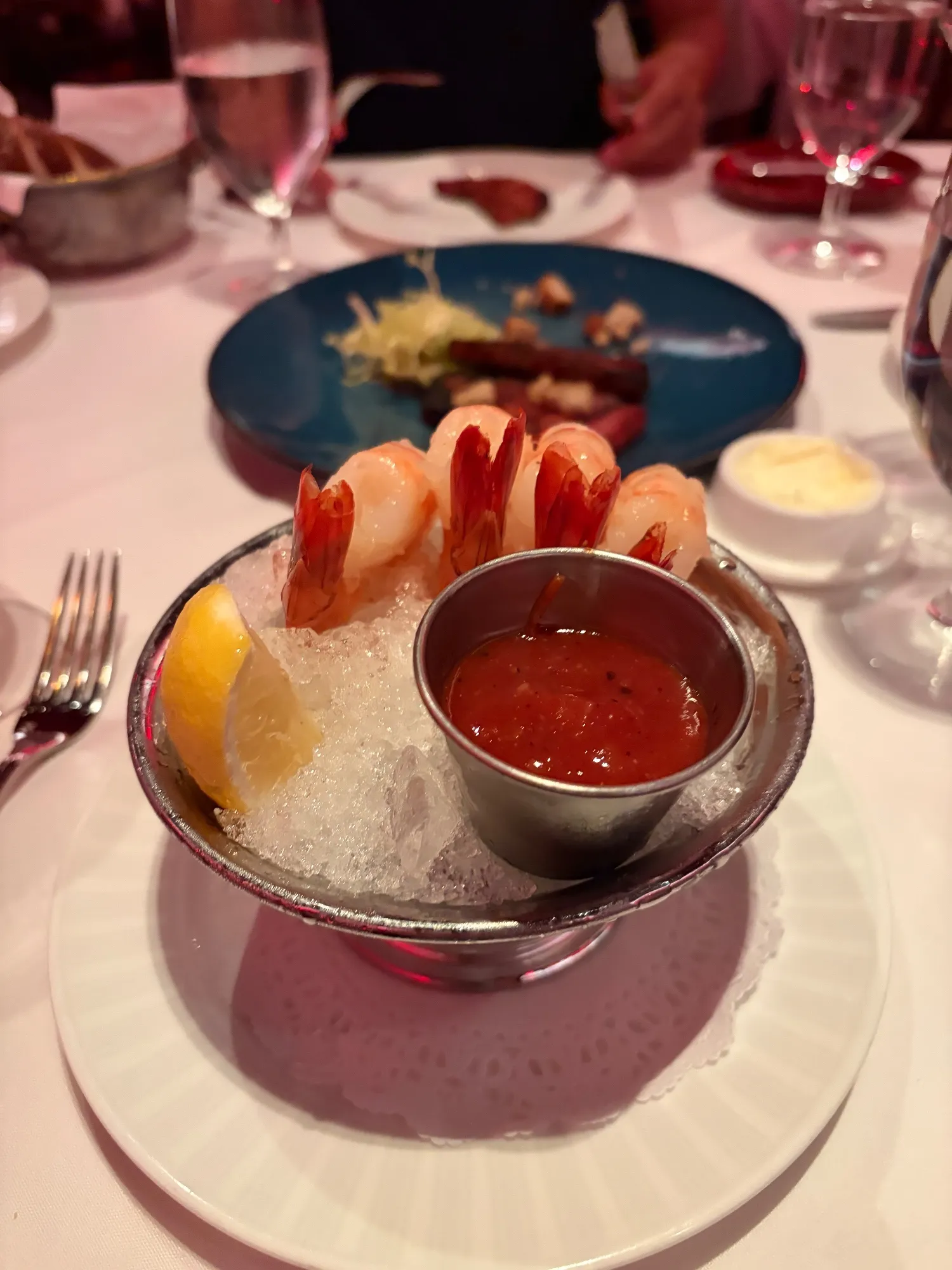 Shrimp arranged over crushed ice around a metal cup of cocktail sauce, with a lemon wedge at the side.