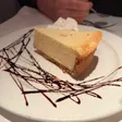 Slice of cheesecake on a white plate with a dollop of whipped cream and a chocolate drizzle pattern.