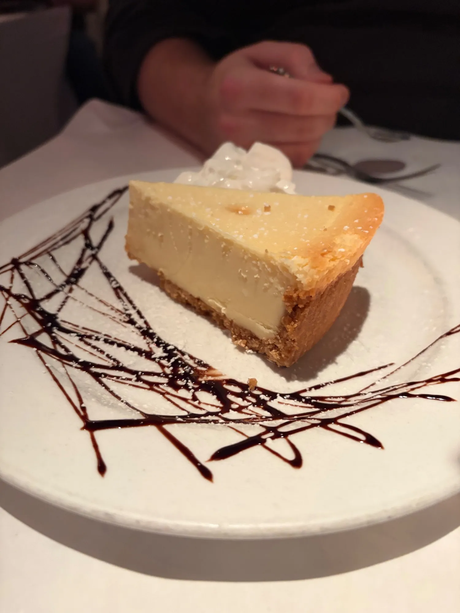 Slice of cheesecake on a white plate with a dollop of whipped cream and a chocolate drizzle pattern.