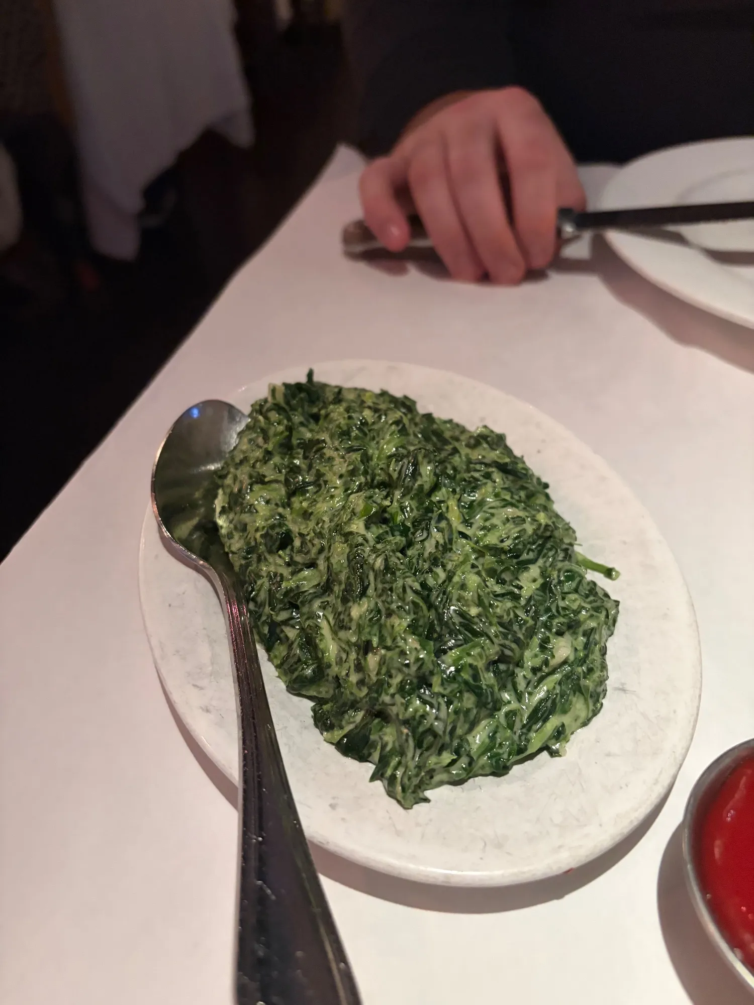 Oval white dish of creamed spinach with a serving spoon; a dish of red sauce at the lower right.