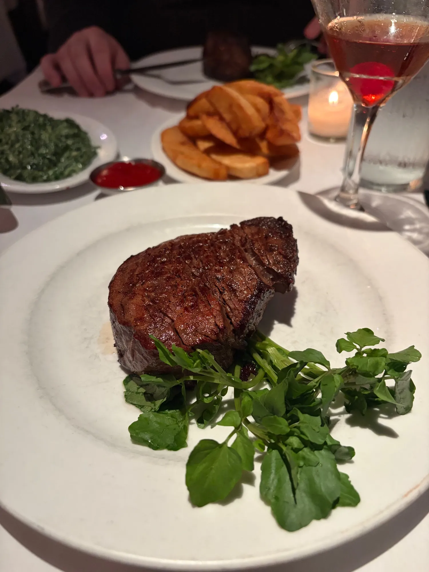 Round filet on a white plate with watercress, a side of fries, creamed spinach, red sauce and a stemmed drink in the background.
