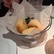 Wire basket lined with paper holding three bread rolls and packets of butter, with a water glass behind.