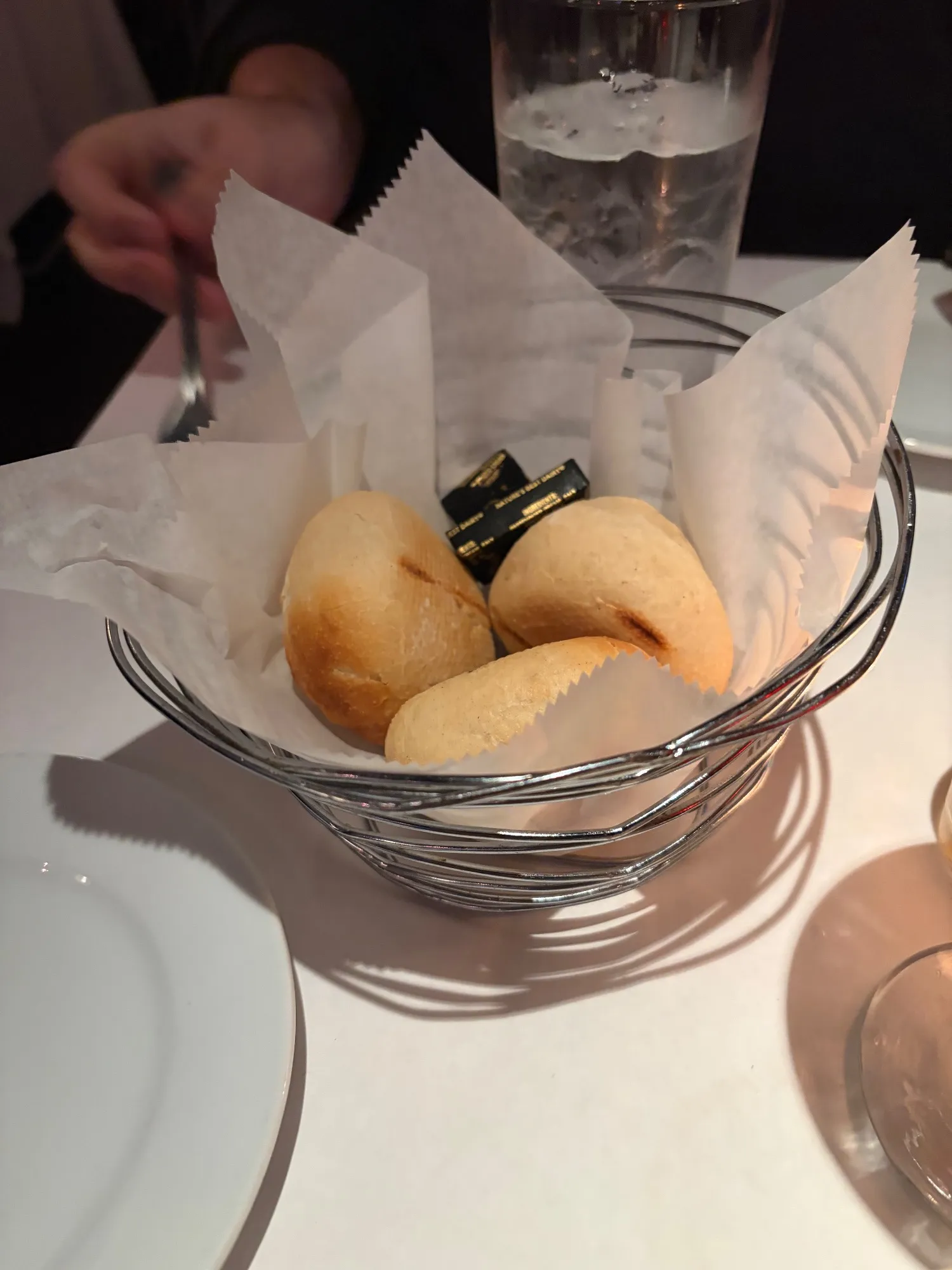 Wire basket lined with paper holding three bread rolls and packets of butter, with a water glass behind.