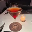 A dark cocktail in a martini glass with a red cherry at the bottom, beside a lit votive candle on a white tablecloth.