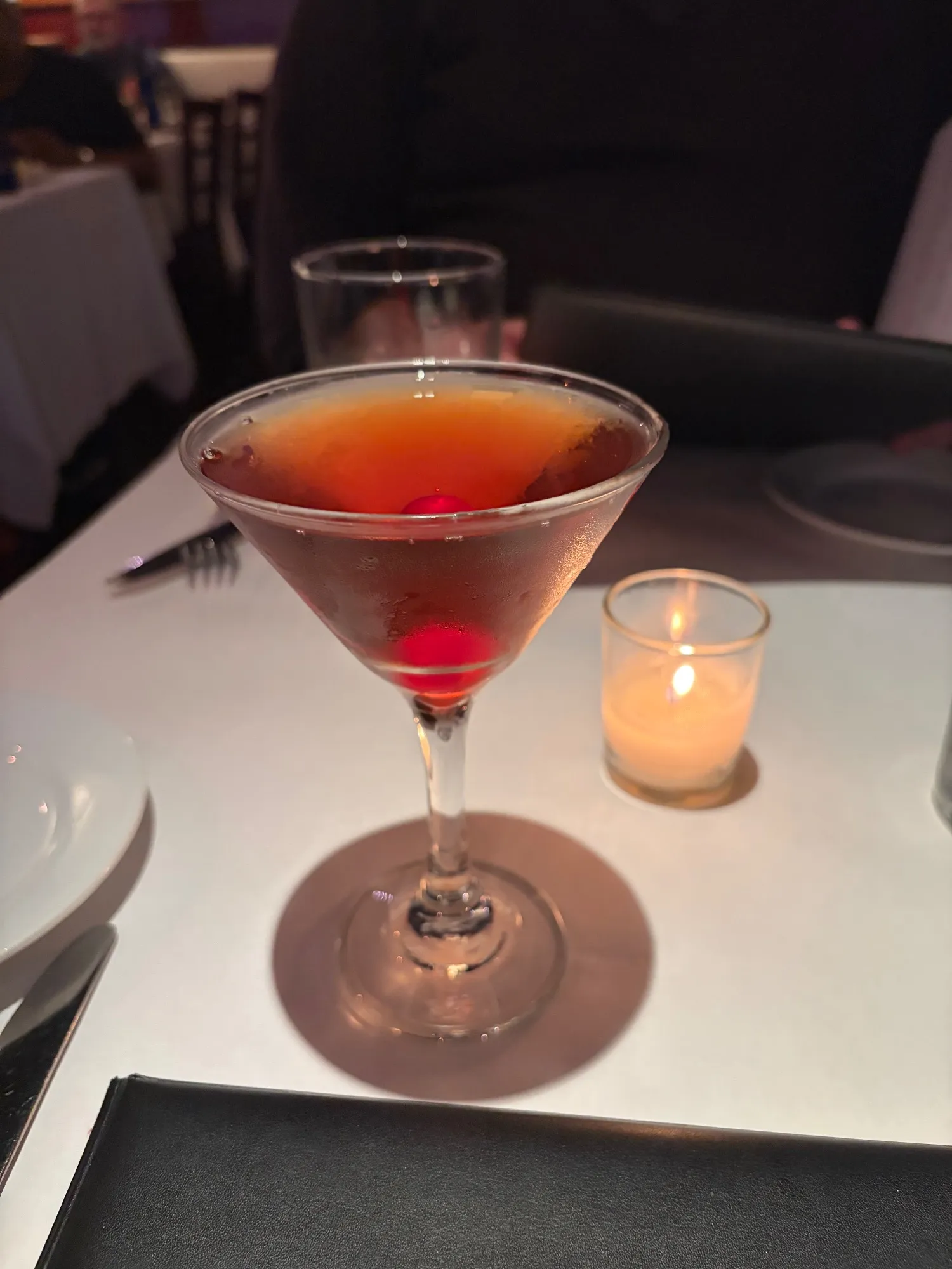 A dark cocktail in a martini glass with a red cherry at the bottom, beside a lit votive candle on a white tablecloth.