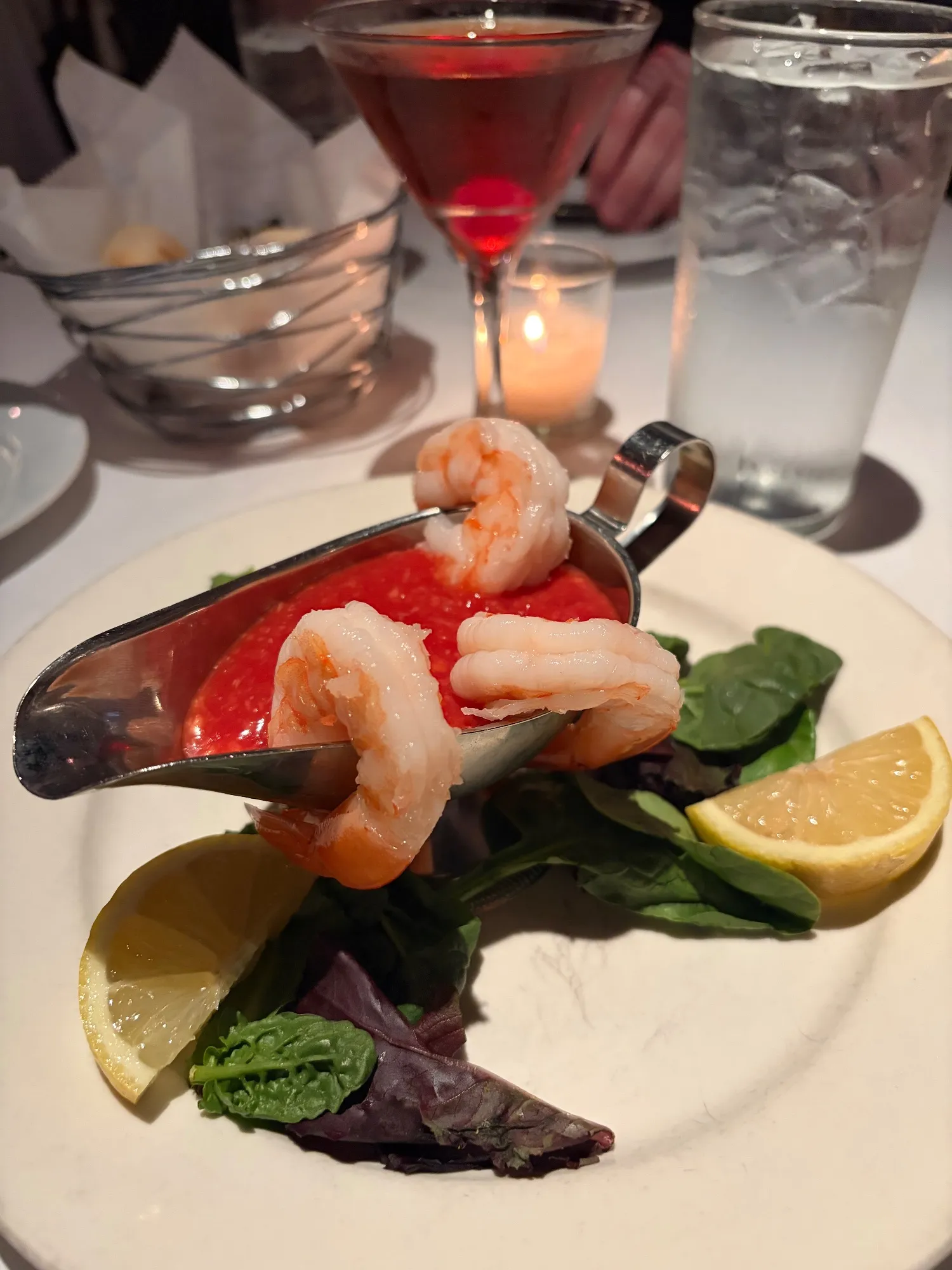 Three shrimp draped over a metal gravy boat of red cocktail sauce, set on greens with lemon wedges; drinks and bread behind.