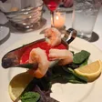 Three shrimp draped over a metal gravy boat of red cocktail sauce, set on greens with lemon wedges; drinks and bread behind.